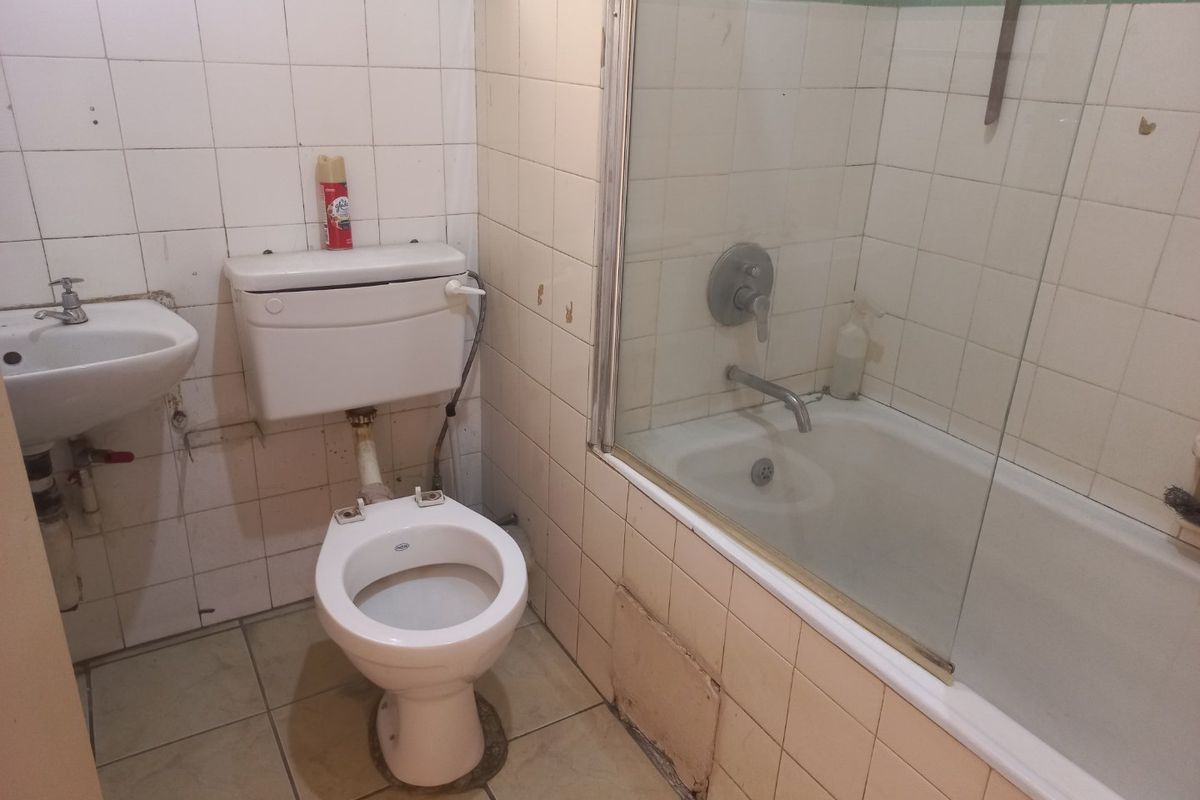 A dated bathroom featuring a bathtub with a glass screen, wall-mounted basin, and toilet, requiring significant renovation and cosmetic updates to modernize the space.