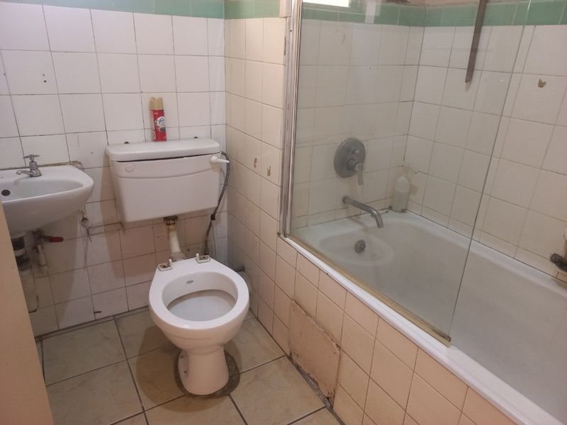 A dated bathroom featuring a bathtub with a glass screen, wall-mounted basin, and toilet, requiring significant renovation and cosmetic updates to modernize the space.