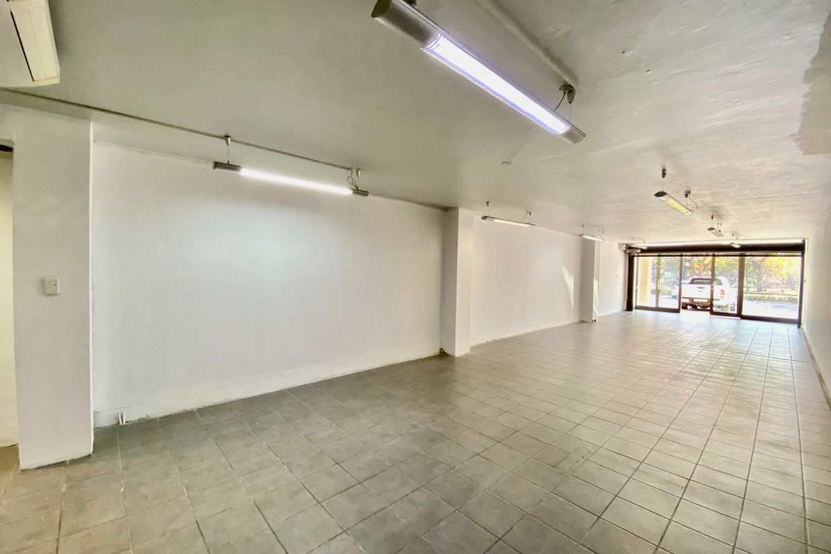 An empty, open-plan commercial space featuring tiled flooring, fluorescent lighting, and large sliding glass doors providing natural light and street access.