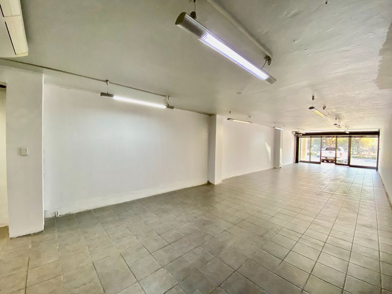 An empty, open-plan commercial space featuring tiled flooring, fluorescent lighting, and large sliding glass doors providing natural light and street access.