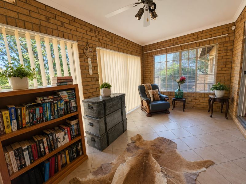 A sunlit living area featuring exposed brick walls, neutral ceramic floor tiles, and large windows with vertical blinds.
