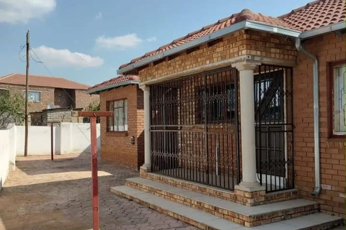 A rear exterior view of a face brick house featuring a covered patio area with security gates and a paved yard.