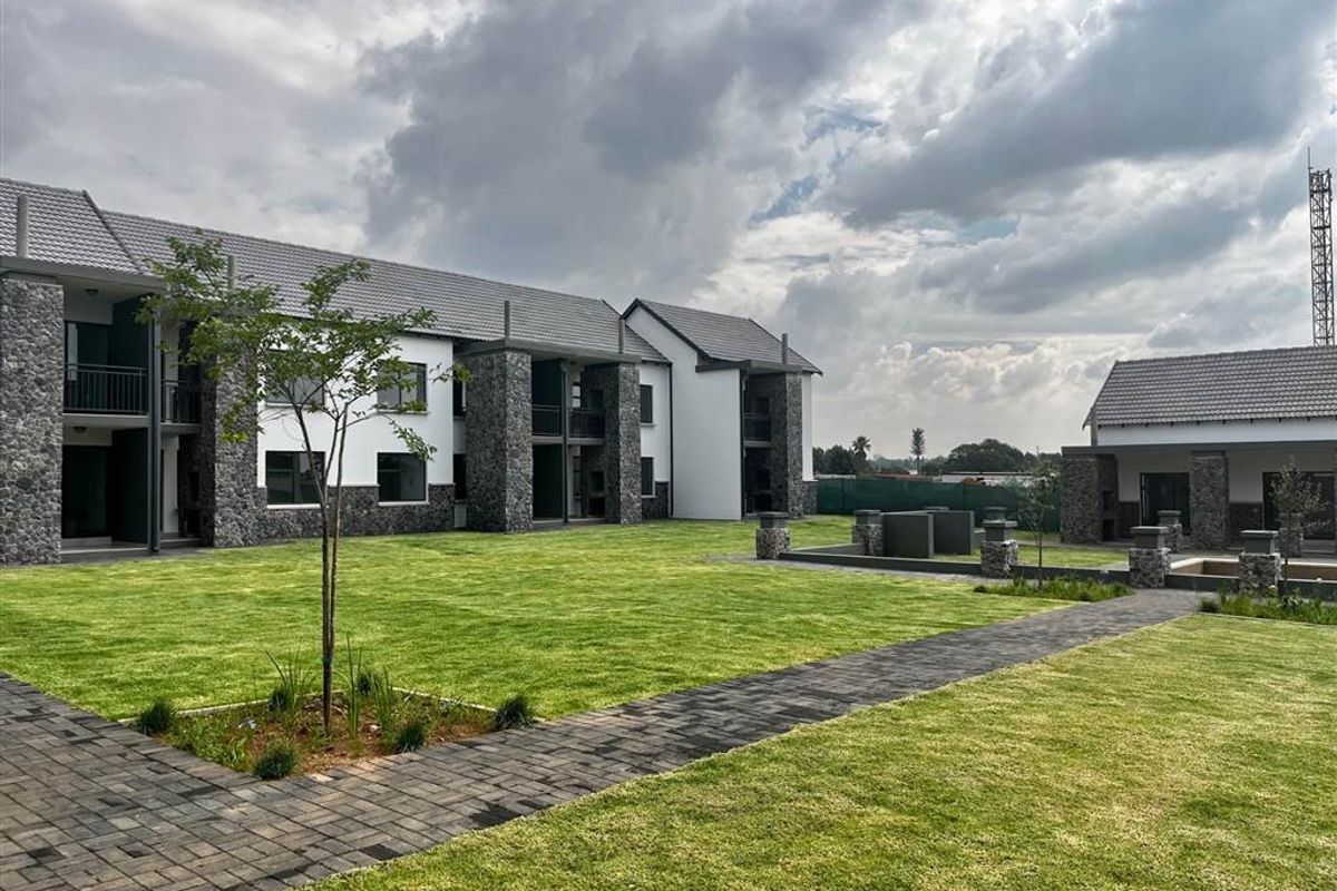 A well-maintained apartment complex exterior featuring stone-clad facades, tiled roofs, manicured lawns, and paved walkways with communal outdoor braai facilities.