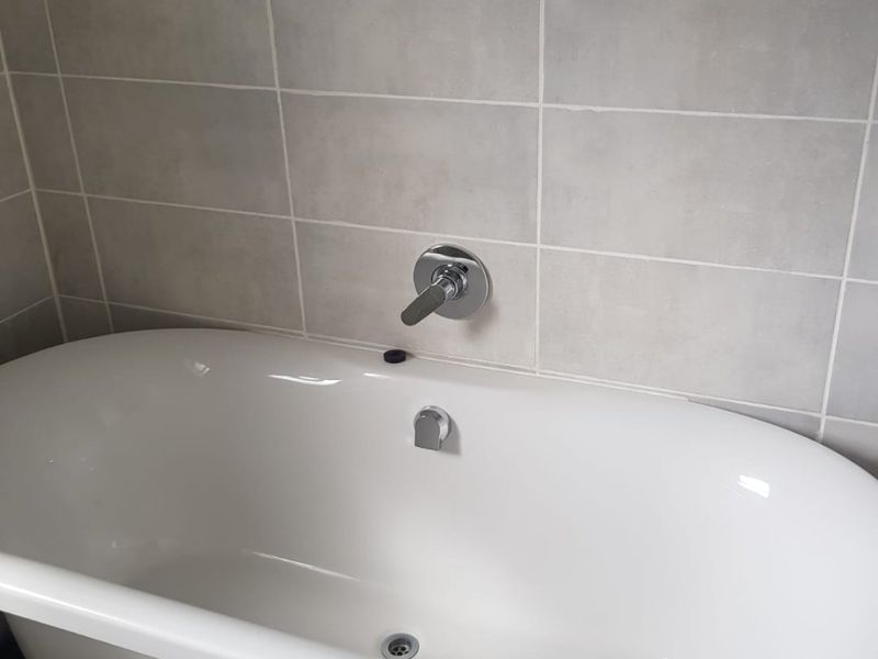 A modern bathroom featuring a clean white freestanding bathtub with wall-mounted chrome mixer tap and neutral grey wall tiling.