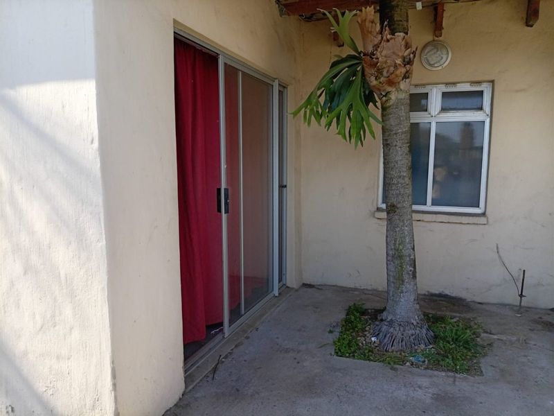 A covered patio area featuring a sliding glass door, concrete flooring, and an exterior wall light, requiring maintenance and cosmetic updates.