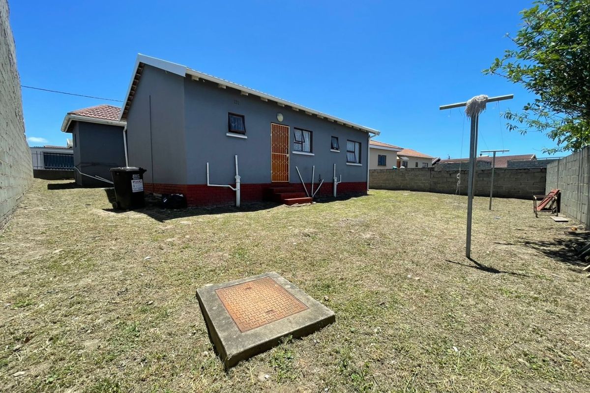 Rear view of the property showing a grey painted exterior, red brick base, security gate, and a spacious backyard with a concrete boundary wall.