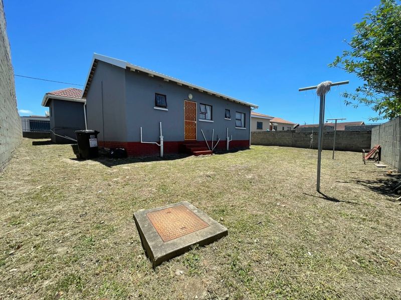 Rear view of the property showing a grey painted exterior, red brick base, security gate, and a spacious backyard with a concrete boundary wall.