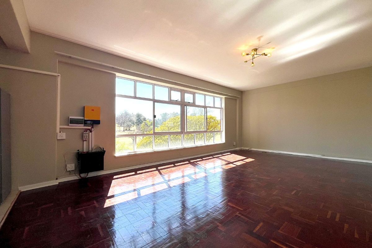 Spacious, sunlit living area featuring polished parquet flooring, large windows with natural views, and a wall-mounted solar inverter system.