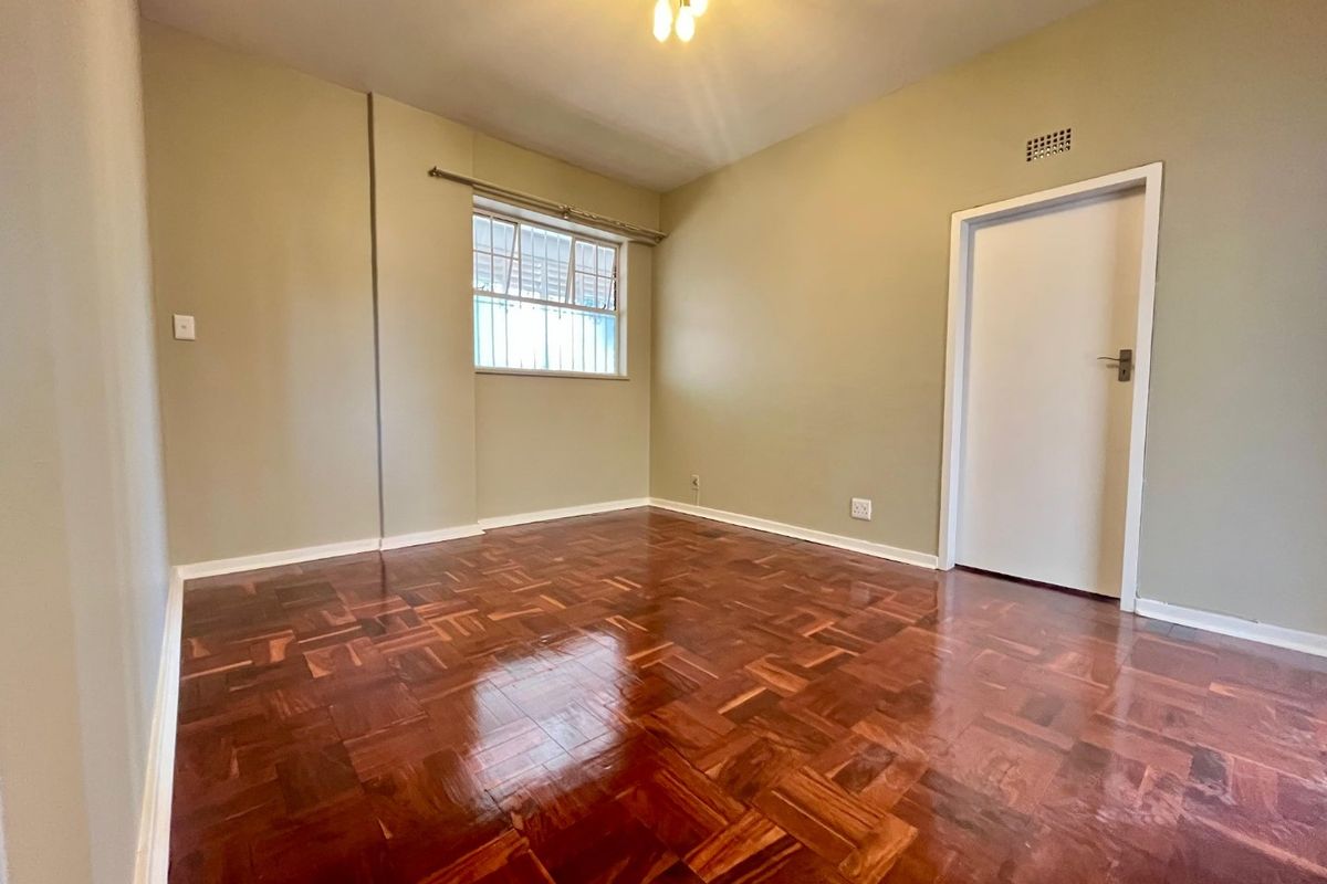 A bright, empty bedroom featuring polished parquet flooring, neutral wall tones, and a window fitted with security bars.