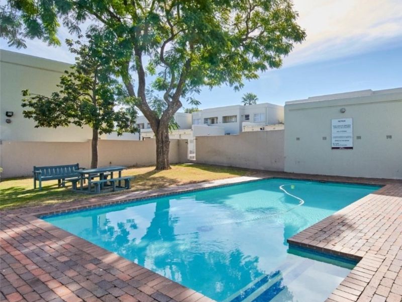 Communal swimming pool area featuring brick paving, a surrounding boundary wall, and mature trees within the apartment complex grounds.