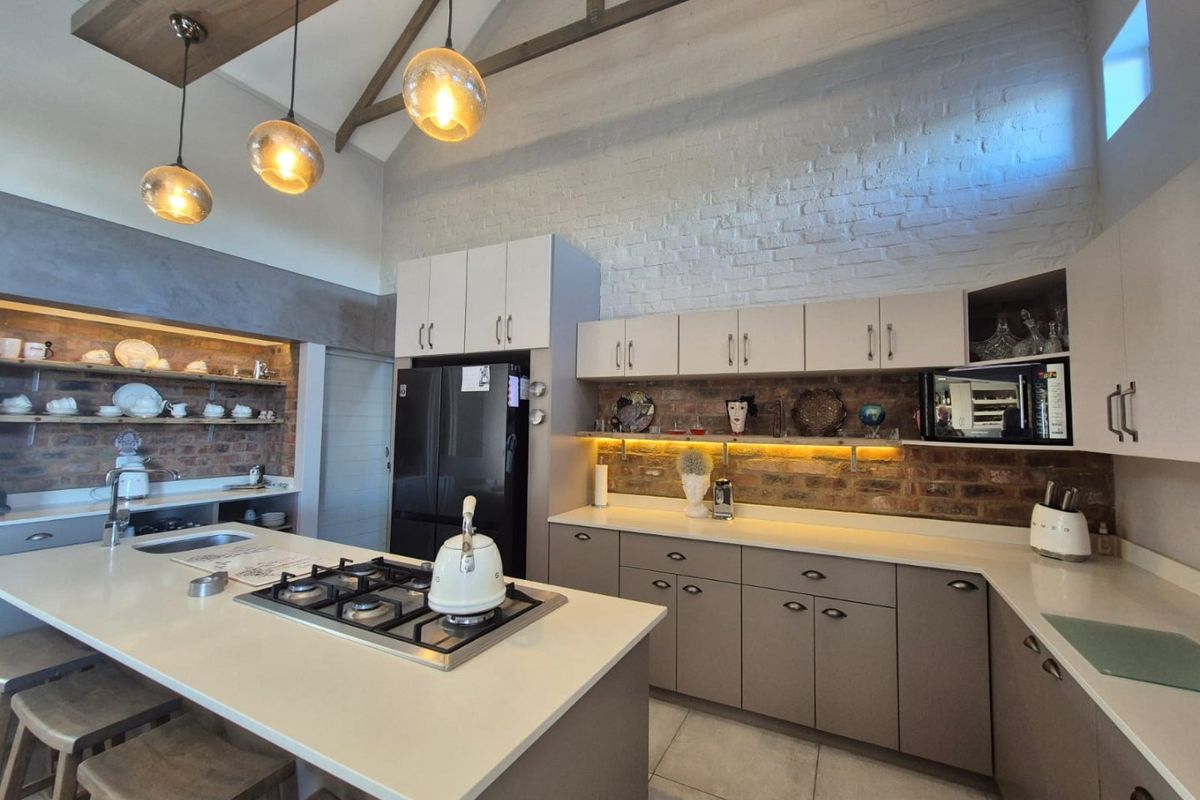 Modern kitchen featuring exposed brick walls, vaulted ceilings with timber beams, a central island with gas cooktop, and high-quality cabinetry with integrated lighting.