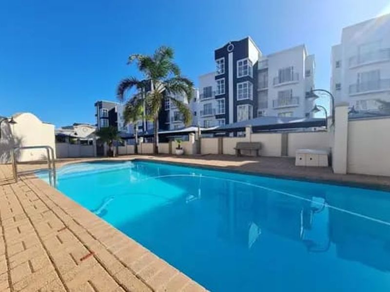 A well-maintained communal swimming pool area featuring a paved deck, pool ladder, and surrounding boundary walls within an apartment complex.