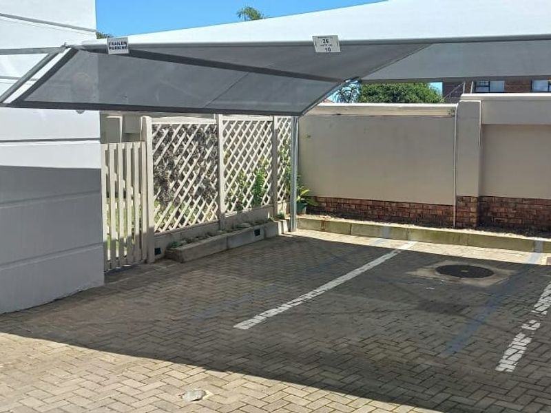 A paved outdoor parking area featuring a fixed shade sail structure, enclosed by a lattice fence and perimeter boundary wall.