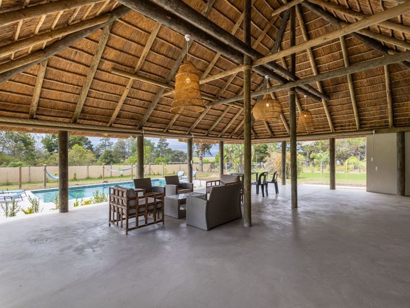 Spacious covered outdoor patio featuring a traditional thatched roof, exposed timber support poles, and concrete flooring, overlooking a private swimming pool and garden.