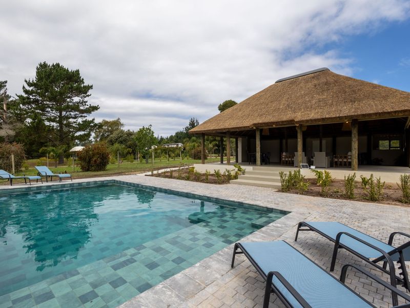 A well-maintained outdoor pool area featuring a tiled swimming pool, paved surround, and a covered patio with a traditional thatched roof structure.