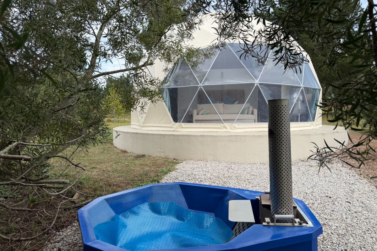 A unique geodesic dome glamping unit set on a concrete base, featuring an adjacent wood-fired hot tub in a natural farm setting.