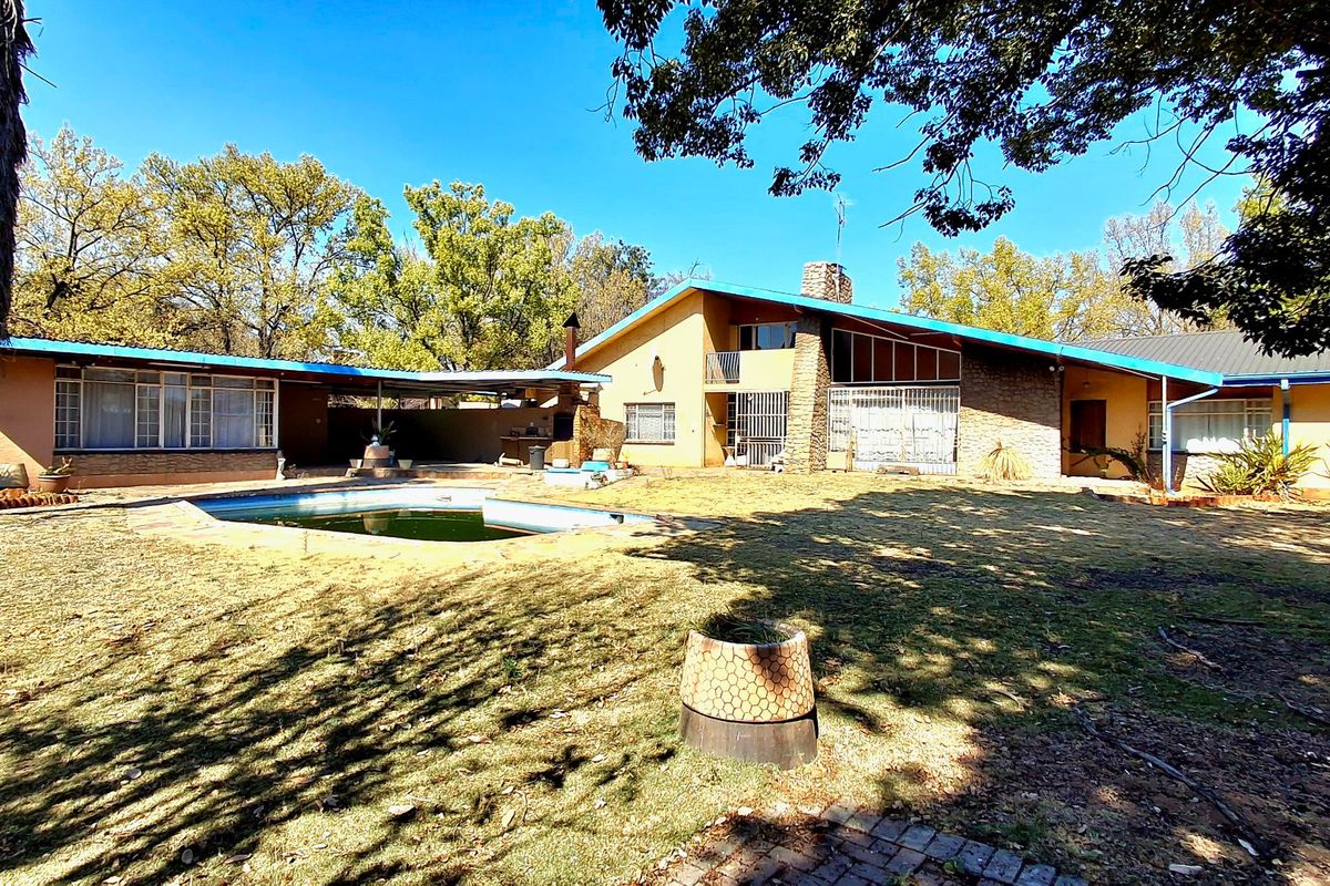 Rear view of a mid-century style house featuring a swimming pool, stone chimney, and covered patio area set within a large garden.
