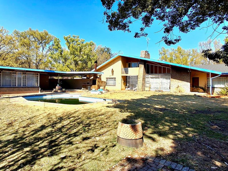 Rear view of a mid-century style house featuring a swimming pool, stone chimney, and covered patio area set within a large garden.