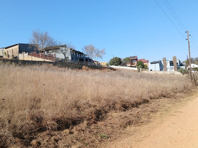 A vacant residential plot featuring natural dry grass vegetation, accessible via a dirt road with established neighboring properties visible in the background.