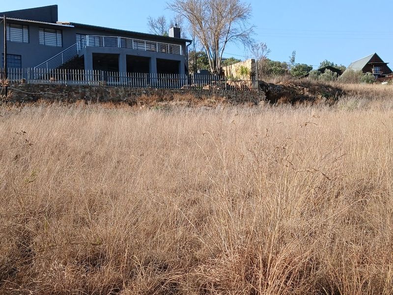 A view of a vacant residential plot featuring dry grass, with a stone retaining wall and an elevated house structure in the background.