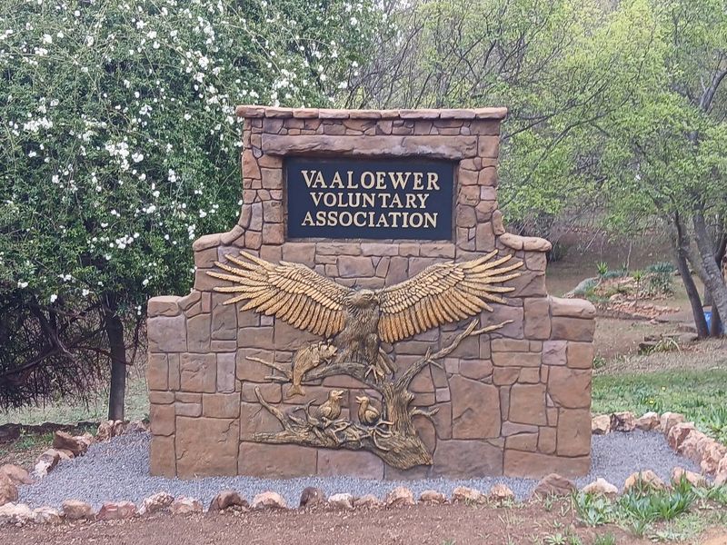 A decorative stone masonry entrance sign for the Vaaloewer Voluntary Association, featuring a detailed eagle relief set within a landscaped garden area.