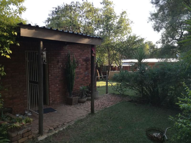 A rear view of the property featuring an exposed brick exterior, a covered patio area with brick paving, and a surrounding garden space.