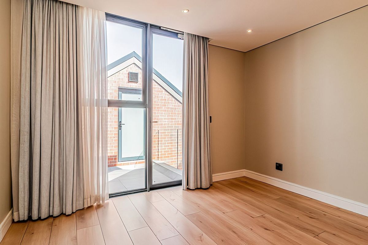 A bright, modern bedroom featuring timber flooring, recessed downlights, and a large sliding glass door providing access to an outdoor balcony area.