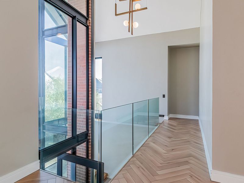 A bright, modern hallway featuring high ceilings, herringbone timber flooring, a sleek glass balustrade, and striking exposed brickwork with expansive floor-to-ceiling windows.