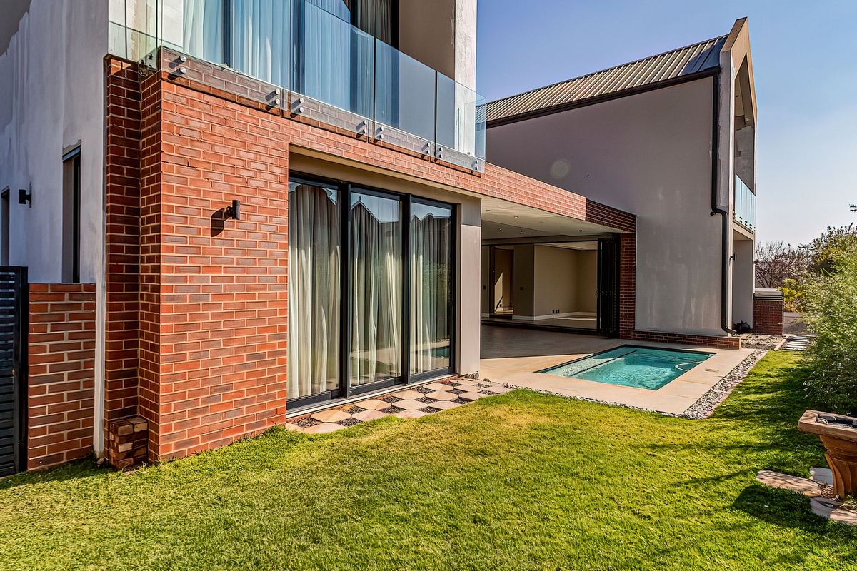 Modern rear exterior featuring a brick facade, covered patio, glass balustrade balcony, and a small swimming pool set within a landscaped garden.