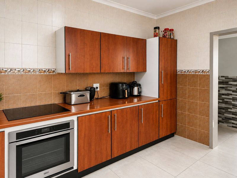 A functional kitchen featuring wood-finish cabinetry, a built-in oven with electric hob, and neutral tiled walls and flooring.