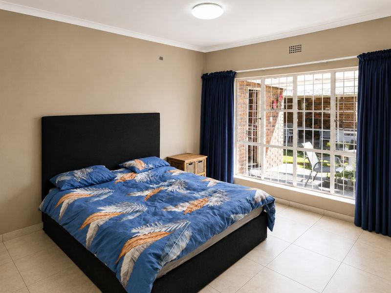 A well-maintained bedroom featuring neutral wall tones, tiled flooring, and a large window with security bars providing natural light and garden views.