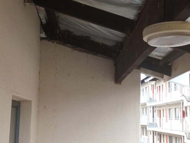 A covered balcony area featuring timber beams and a metal railing, showing signs of wear and water staining on the exterior wall.