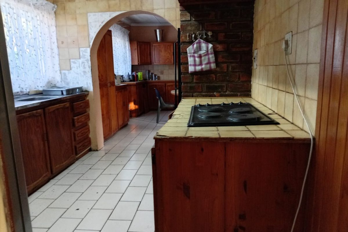 A dated kitchen featuring wooden cabinetry, tiled countertops, an electric hob, and a brick feature wall, connected to an adjacent scullery area via an archway.