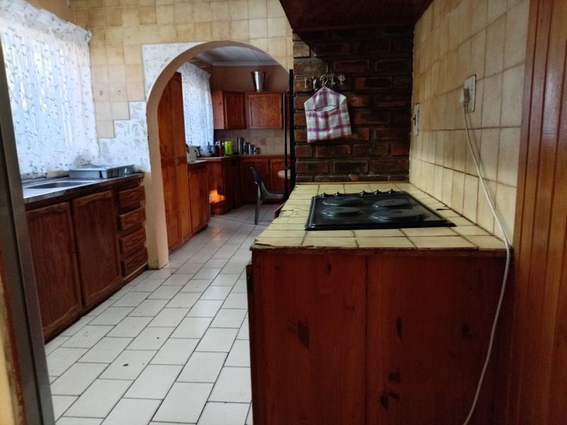 A dated kitchen featuring wooden cabinetry, tiled countertops, an electric hob, and a brick feature wall, connected to an adjacent scullery area via an archway.