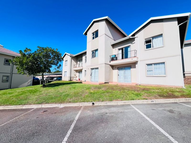 Exterior view of a multi-story apartment complex featuring paved parking bays, landscaped lawn, and balcony access with security gates.