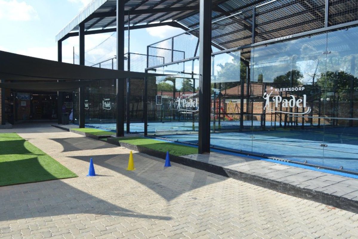 Modern outdoor padel court facility featuring professional glass enclosures, steel structural framing, and paved walkways with artificial turf accents.