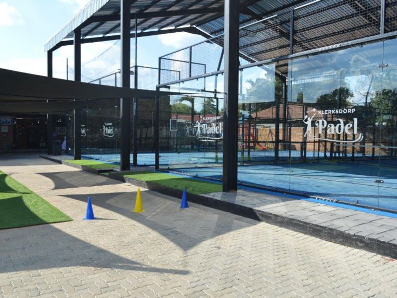 Modern outdoor padel court facility featuring professional glass enclosures, steel structural framing, and paved walkways with artificial turf accents.