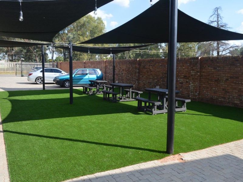 Outdoor commercial patio area featuring artificial turf, shade sails supported by steel poles, and a brick boundary wall.