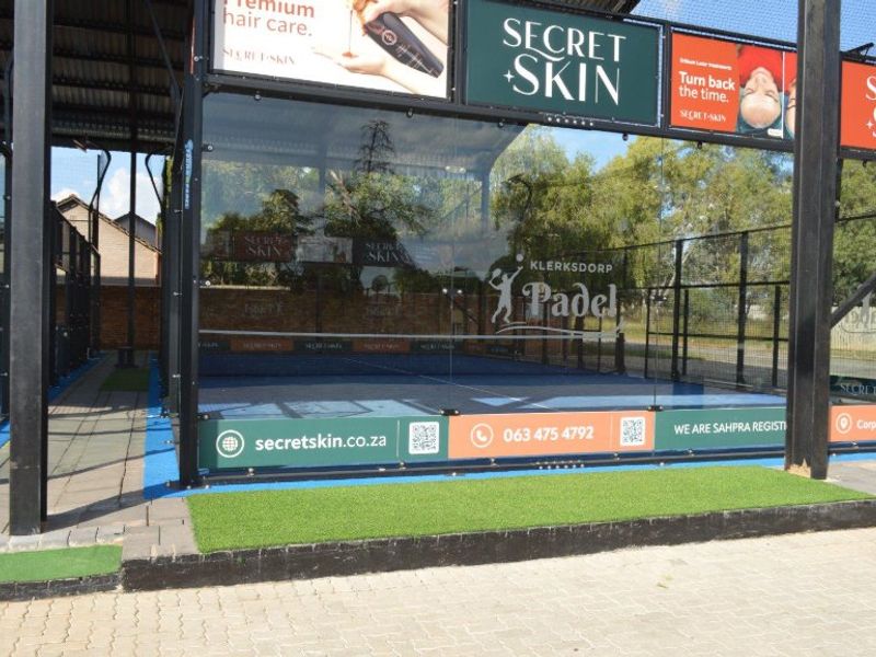 A well-maintained outdoor padel court featuring glass perimeter walls, synthetic turf, and integrated commercial advertising signage.