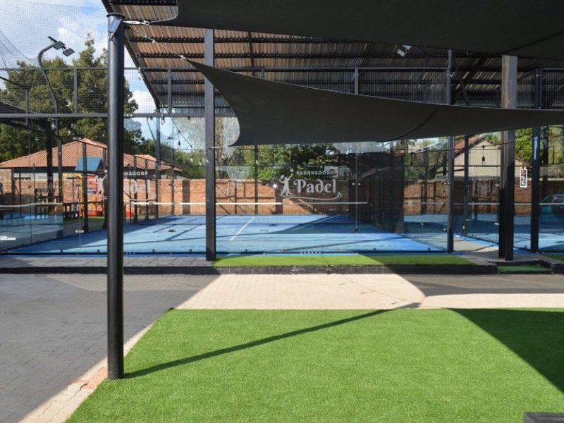 A well-maintained commercial padel court facility featuring glass walls, metal roofing, artificial turf surfacing, and integrated shade sails for outdoor play.