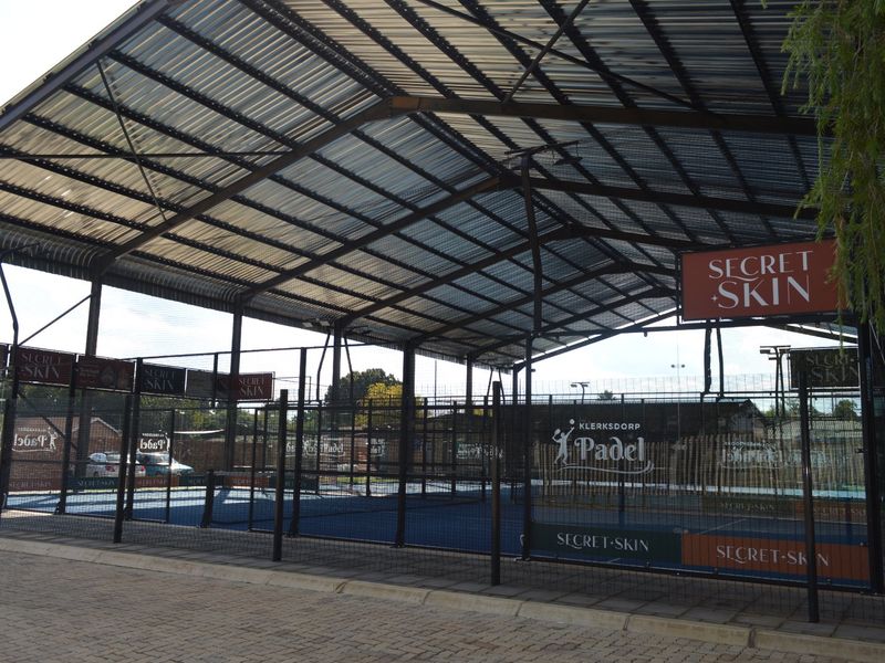 A well-maintained outdoor padel court facility featuring a large protective metal roof structure, steel fencing, and professional court surfacing.