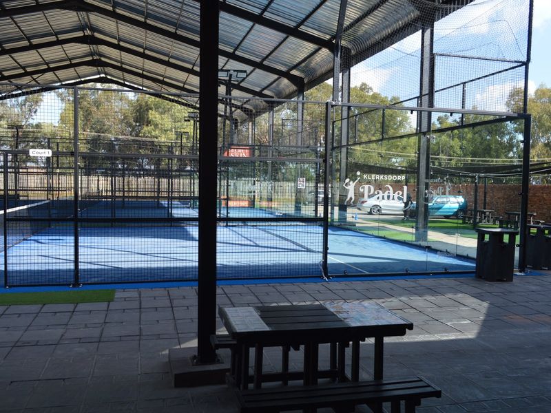 A well-maintained covered padel court facility featuring professional glass walls, metal mesh fencing, and paved spectator area under a steel roof structure.