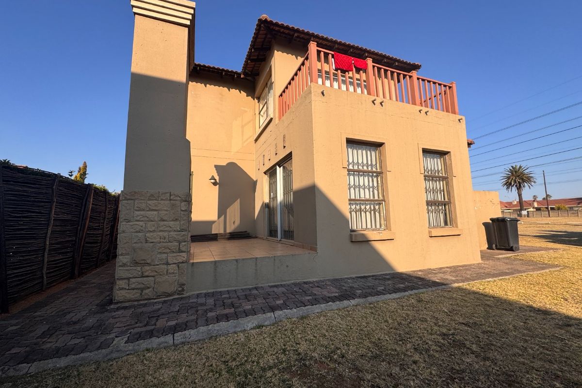Rear view of a double-story house featuring a tiled patio, upper-level balcony with wooden railing, stone cladding accents, and paved garden pathways.