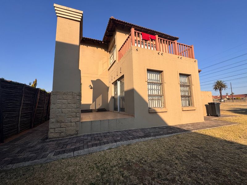 Rear view of a double-story house featuring a tiled patio, upper-level balcony with wooden railing, stone cladding accents, and paved garden pathways.