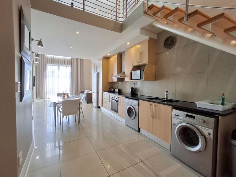 Modern open-plan kitchen and dining area featuring sleek cabinetry, integrated appliances, tiled flooring, and a staircase leading to an upper level.