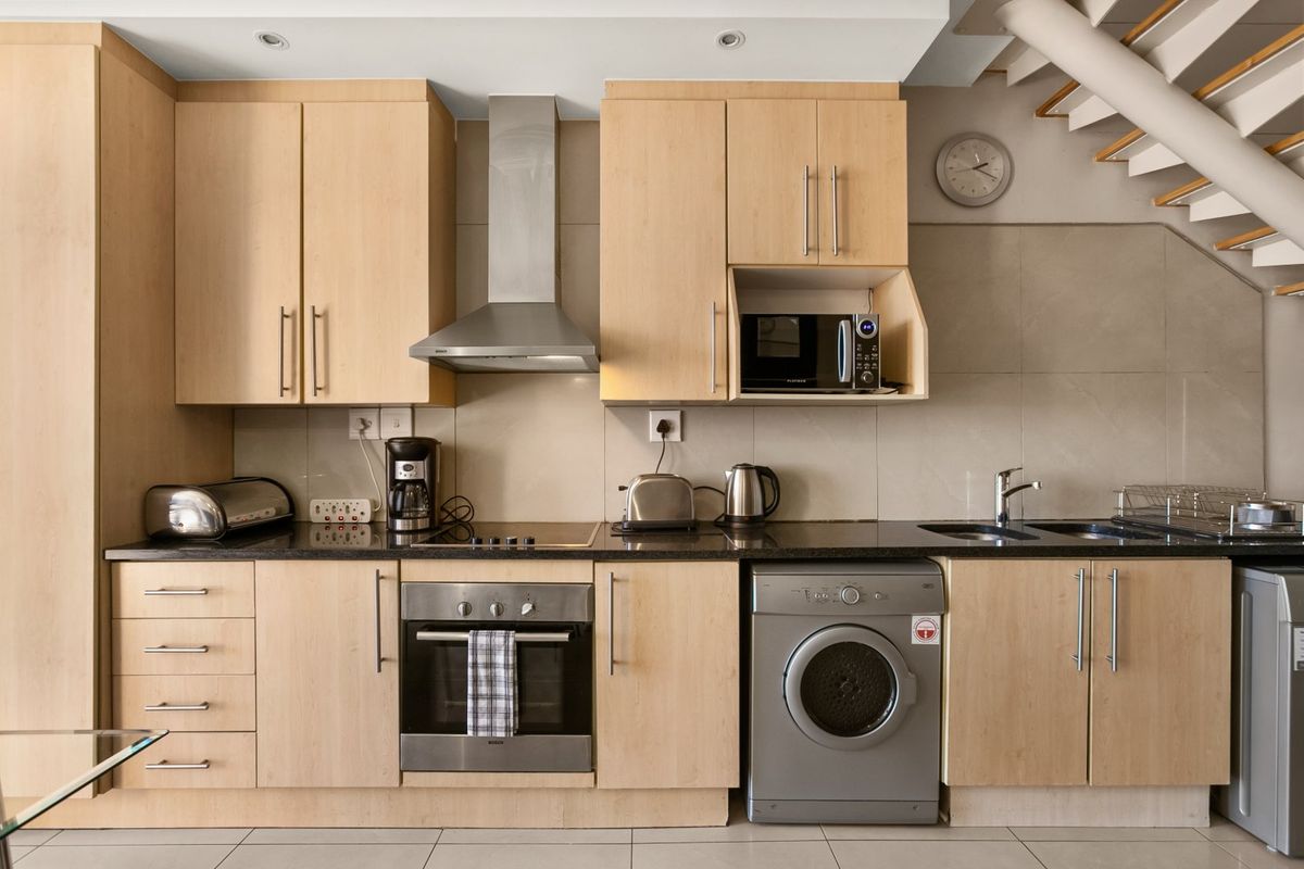 Modern kitchen featuring light-wood cabinetry, granite countertops, integrated stainless steel appliances, and a tiled backsplash, conveniently located beneath a staircase.