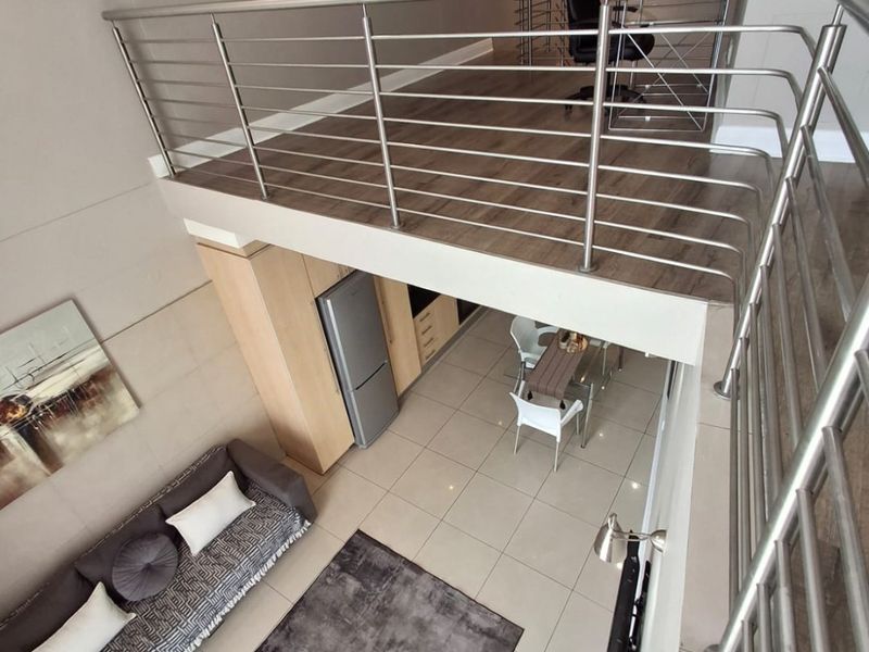 A modern open-plan apartment featuring a mezzanine level with stainless steel balustrades, overlooking a living and dining area with tiled floors and built-in cabinetry.