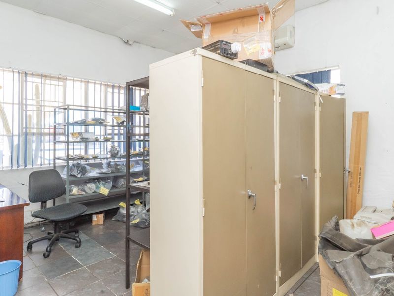 Functional industrial office space featuring tiled flooring, security bars on windows, and overhead fluorescent lighting, suitable for administrative or storage use.