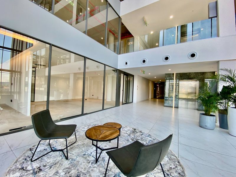 Modern commercial building entrance lobby featuring double-volume ceilings, glass-partitioned office spaces, marble-look flooring, and a sleek elevator access point.