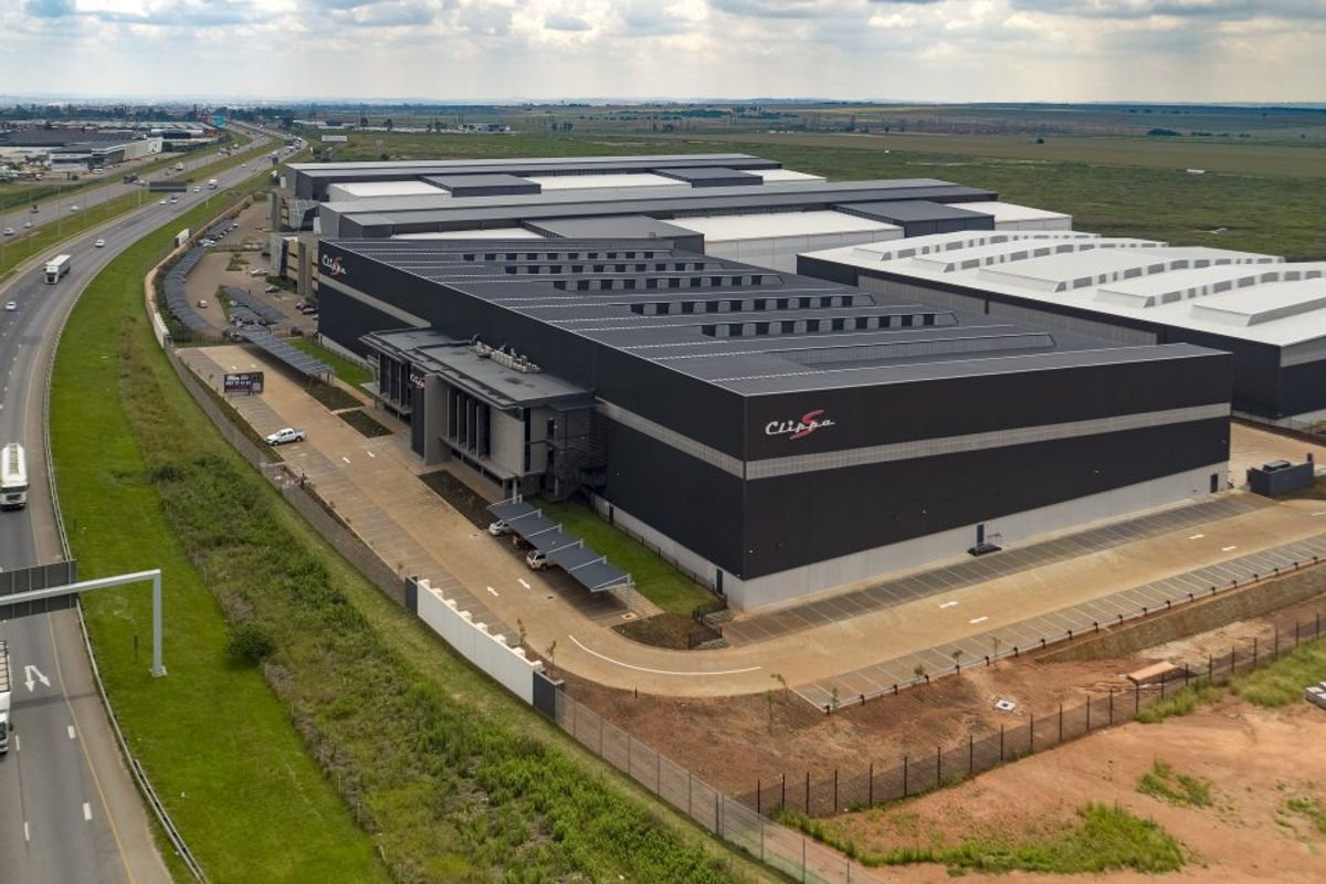 Aerial view of a large-scale industrial warehouse facility featuring extensive roofing, paved parking areas, and secure perimeter fencing adjacent to a major highway.